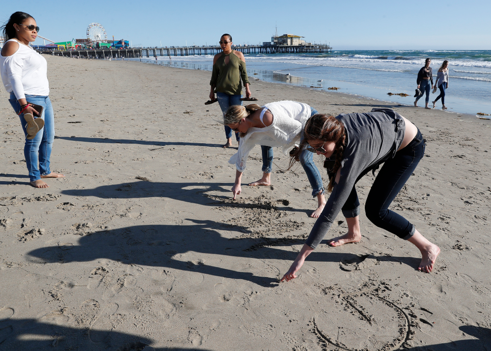 Iowa Hawkeyes guard Tania Davis (11), guard Makenzie Meyer (3), forward Megan Gustafson (10), and guard Alexis Sevillian (5) write Go Hawkeyes in the sand as they explore the beach Thursday, March 15, 2018 in Santa Monica. (Brian Ray/hawkeyesports.com)